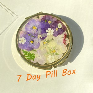 Könnte beinhalten: Eine runde Pillendose aus Gold mit einem Scharnierdeckel. Der Deckel ist mit gepressten Blumen in Violett-, Weiß- und Gelbtönen verziert. Der Text "7 Day Pill Box" ist in Orange unter der Dose aufgedruckt.