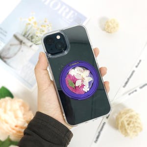 May include: A clear phone case with a black back and a purple ring holder. The ring holder has a clear window with pressed flowers inside.