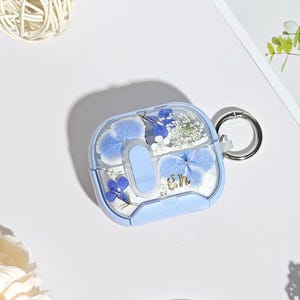Può includere: Una custodia azzurra per AirPods con parte superiore trasparente, con fiori blu pressati e dettagli bianchi. La custodia ha un portachiavi argentato e le iniziali dorate "gh". Progettata per proteggere e personalizzare gli AirPods.