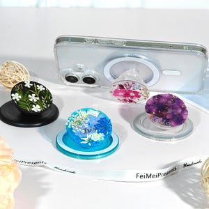 May include: A collection of decorative phone grips with floral designs. One grip features white flowers on a black background, another has blue flowers, and a third showcases purple flowers. A clear phone case with a grip attached is also present.