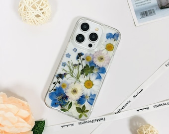 May include: Clear phone case with embedded dried flowers. The case features a variety of blue, white, and yellow flowers, including daisies and forget-me-nots. The case has a clear border and a camera cutout.