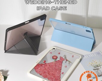 Op de afbeelding: Drie iPad-hoesjes, elk in een andere kleur, worden getoond. Eén is zwart, één is lichtblauw en de derde is wit met een ontwerp van geperste bloemen. De tekst "HAND-PAINTED PRESSED FLOWER WEDDING-THEMED IPAD CASE" staat bovenaan.