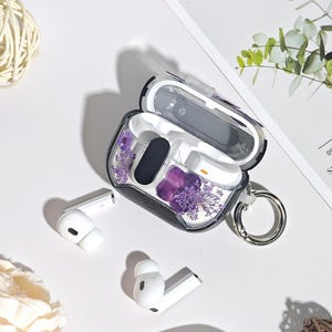 Può includere: Una custodia protettiva trasparente per auricolari wireless, con fiori secchi viola incorporati. La custodia è aperta, rivelando gli auricolari bianchi all'interno. È attaccato un portachiavi argentato.