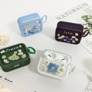 May include: Four AirPod cases in various colors: light blue, dark purple, green, and white. Each case features a floral design with the names "Ruby", "Isla", and "Freya" printed on them. The white case is open, revealing white earbuds.