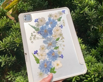 May include: A white tablet case with a clear back panel featuring pressed blue and white flowers. The case has a camera cutout and a white border.