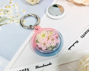 May include: A clear, round keychain with a pink and blue gradient border, filled with dried flowers. The keychain has a silver metal clasp and a small, white, circular device is visible in the background.
