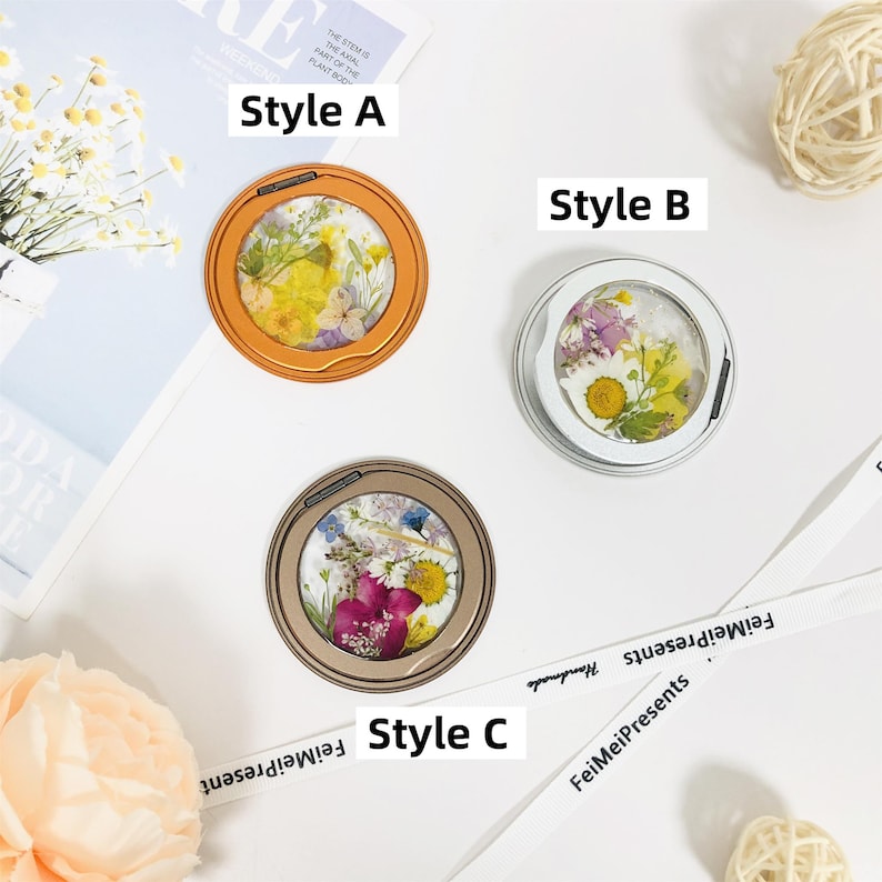 May include: Three round, decorative phone grips with different styles of pressed flowers. The phone grips are in orange, white, and gold frames. The text "Style A", "Style B", and "Style C" are written below each phone grip.
