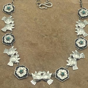 May include: A silver necklace featuring alternating links of stylized birds and floral motifs. Each flower link contains a small, round turquoise stone at its center. The necklace includes a chain with a hook clasp and an adjustable extender link.