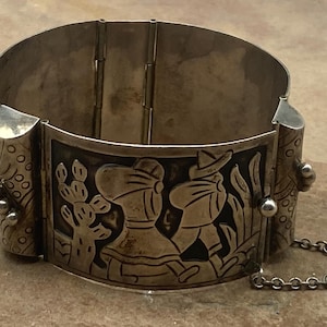 May include: A wide silver hinged cuff bracelet featuring a central panel with a relief design of two figures walking past a cactus. The side panels have a textured pattern with small silver spheres, and a thin safety chain is attached to the side.
