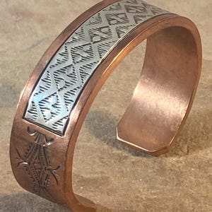 Peut inclure: Un bracelet manchette en cuivre doté d'une incrustation centrale argentée avec un motif en losange répétitif. La bande de cuivre est finie avec des détails géométriques estampés sur les côtés, contrastant le métal chaud et l'argent.
