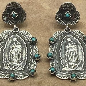 May include: A pair of silver dangle earrings featuring an embossed image of the Virgin of Guadalupe. Each earring is accented with small, round turquoise stones set along the scalloped edges and a single turquoise stone in the top post.