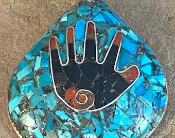 Puede incluir: Colgante en forma de lágrima con una fianza plateada. El colgante presenta un mosaico de piedras turquesas que rodea un diseño de mano negra con acentos rojos. La mano está delineada en plata.