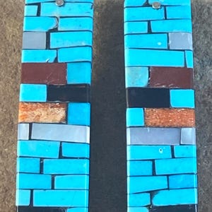 May include: A pair of rectangular turquoise earrings with a mosaic design. The earrings feature a mix of turquoise, brown, black, and gray pieces, arranged in a brick-like pattern. Each earring has a silver-colored hook.