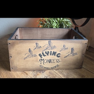 May include: A rectangular wooden crate with the words "Flying Monkeys Air Mail Service" printed on the front. The crate is stained brown with black lettering and illustrations of monkeys with wings. The crate has two handle holes on the sides.