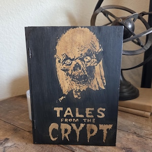 May include: A black wooden sign with the words "TALES FROM THE CRYPT" in a distressed font. The sign features a carved image of a ghoul's face in a light brown color. The sign is propped up on a wooden surface.
