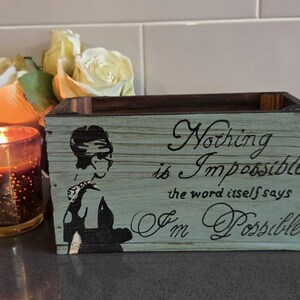 May include: A wooden box with a light blue painted finish and the words "Nothing is Impossible" in black script. A black and white silhouette of a woman is on the side. A lit candle and flowers are next to the box.