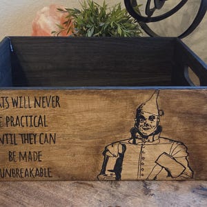 May include: Wooden storage box with a dark stained exterior and blue interior. The box features a quote and an illustration of the Tin Man from The Wizard of Oz. The quote reads: "Hearts will never be practical until they can be made unbreakable."