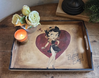 Puede incluir: Bandeja de madera con un diseño de Betty Boop pintado a mano. La bandeja presenta un borde negro, asas y un fondo en forma de corazón. También hay una vela encendida y un pequeño jarrón con rosas.