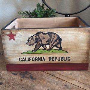 May include: Wooden box with the California Republic emblem. The box features a brown bear on green grass, a red star, and the words "CALIFORNIA REPUBLIC" in black. The bottom of the box is painted red.