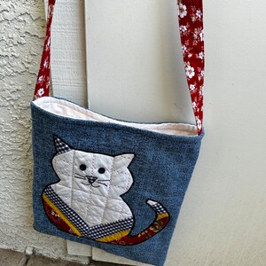 K&ouml;nnte beinhalten: Eine denimblaue Umh&auml;ngetasche mit einem wei&szlig;en, gesteppten Katzen-Applik. Die Katze hat schwarze Augen, eine rosa Nase und einen rot-gelb-schwarz gemusterten Schwanz. Die Tasche hat einen roten Riemen mit wei&szlig;em Blumendruck.
