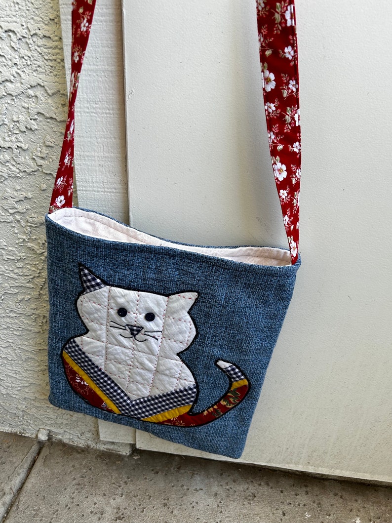 K&ouml;nnte beinhalten: Eine denimblaue Umh&auml;ngetasche mit einem wei&szlig;en, gesteppten Katzen-Applik. Die Katze hat schwarze Augen, eine rosa Nase und einen rot-gelb-schwarz gemusterten Schwanz. Die Tasche hat einen roten Riemen mit wei&szlig;em Blumendruck.