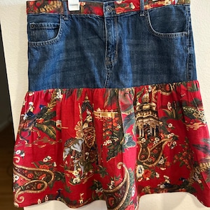 May include: A denim skirt with a red patterned bottom. The skirt features a classic denim top with pockets and a button closure. The bottom is a vibrant red fabric adorned with an intricate design of elephants, temples, and paisley patterns.