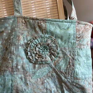May include: A quilted tote bag in shades of green and brown, featuring a large, fabric flower applique. The bag has a floral pattern and is stitched with a grid pattern. The handles are made of the same fabric.