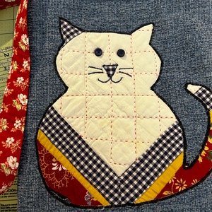 K&ouml;nnte beinhalten: Eine wei&szlig;e, gesteppte Tasche in Katzenform mit schwarzen und wei&szlig;en Karomustern. Die Tasche hat einen roten und wei&szlig;en Blumendruck-Trageriemen und ist aus Jeansstoff.