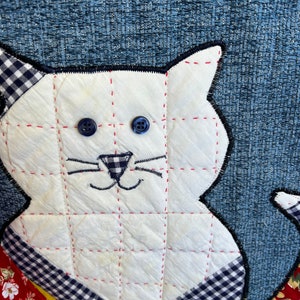 K&ouml;nnte beinhalten: Eine wei&szlig;e gesteppte Katze mit blauen Knopfaugen und einer schwarz-wei&szlig; karierten Nase. Die Katze sitzt auf einem blau-wei&szlig; karierten Stoff mit roten und gelben Akzenten.