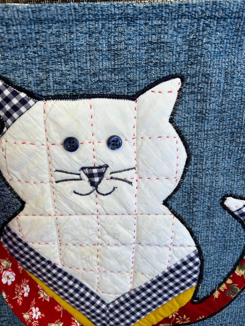 K&ouml;nnte beinhalten: Eine wei&szlig;e gesteppte Katze mit blauen Knopfaugen und einer schwarz-wei&szlig; karierten Nase. Die Katze sitzt auf einem blau-wei&szlig; karierten Stoff mit roten und gelben Akzenten.