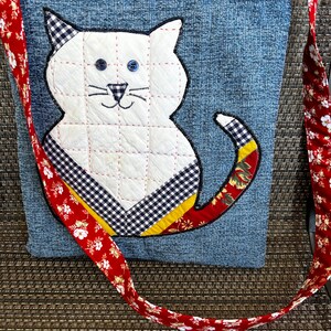 K&ouml;nnte beinhalten: Eine Denim-Tragetasche mit einer wei&szlig;en Katzen-Applikation. Die Katze hat auf dem R&uuml;cken ein schwarz-wei&szlig;es Karomuster und einen roten, gelben und gr&uuml;nen Blumenschwanz. Die Tasche hat einen roten und wei&szlig;en Blumengurt.