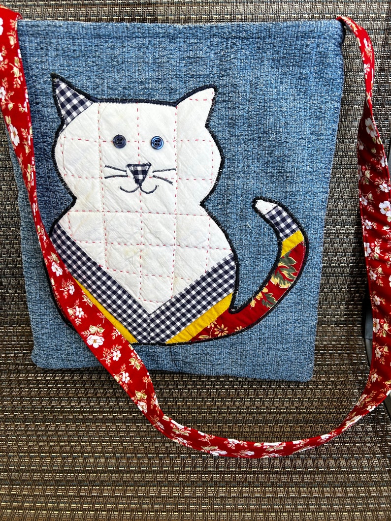 K&ouml;nnte beinhalten: Eine Denim-Tragetasche mit einer wei&szlig;en Katzen-Applikation. Die Katze hat auf dem R&uuml;cken ein schwarz-wei&szlig;es Karomuster und einen roten, gelben und gr&uuml;nen Blumenschwanz. Die Tasche hat einen roten und wei&szlig;en Blumengurt.