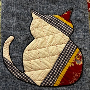 K&ouml;nnte beinhalten: Eine blaue Jeans-Tragetasche mit einer wei&szlig;en gesteppten Katzen-Applikation. Die Katze ist mit rotem, gelbem und schwarzem kariertem Stoff verziert. Die Tasche hat einen roten und wei&szlig;en Blumensatinband-Henkel.