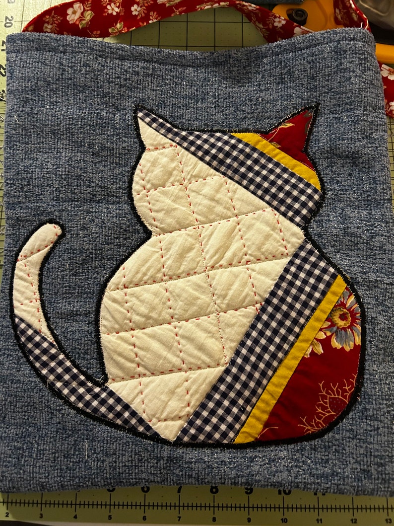 K&ouml;nnte beinhalten: Eine blaue Jeans-Tragetasche mit einer wei&szlig;en gesteppten Katzen-Applikation. Die Katze ist mit rotem, gelbem und schwarzem kariertem Stoff verziert. Die Tasche hat einen roten und wei&szlig;en Blumensatinband-Henkel.
