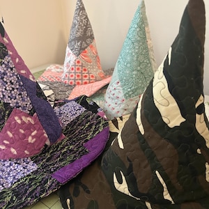 May include: A collection of four quilted cone-shaped hats featuring various fabric patterns. One hat displays a purple floral print, another a camouflage design, a third a light green botanical pattern, and the fourth a grey and coral patchwork.