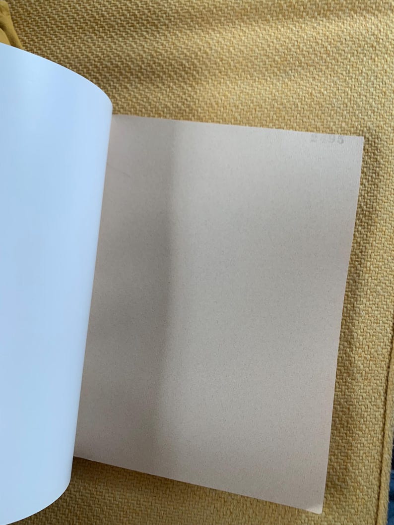 May include: Open book with white and light tan pages. The left side of the book shows the white pages, while the right side displays a light tan page. The book is open on a yellow textured surface.