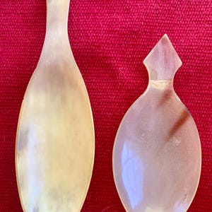 May include: Two smooth, polished horn spoons with natural variations in color. The left spoon is elongated with a rounded handle, while the right spoon is wider and oval-shaped with a pointed, diamond-shaped handle. Both feature translucent, creamy beige and light brown tones.