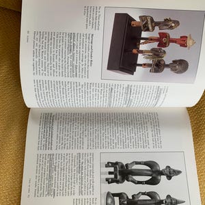 May include: A page from a book featuring images of African art. The top image displays several carved wooden figures on a black stand. The figures are in various colors, including red, brown, and white. The page also contains text.