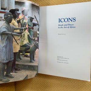 May include: An open book with the title "ICONS Ideals and Power in the Art of Africa" on the right page. The left page shows a photograph of several statues of people in a public space. The statues are in front of a building.