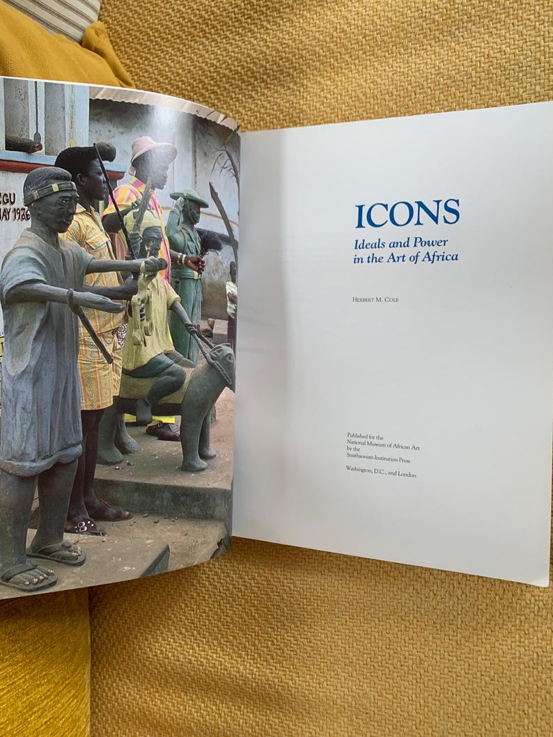 May include: An open book with the title "ICONS Ideals and Power in the Art of Africa" on the right page. The left page shows a photograph of several statues of people in a public space. The statues are in front of a building.
