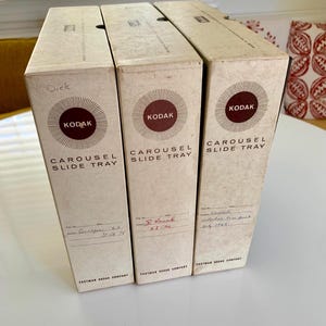 May include: Three vintage Kodak Carousel Slide Tray boxes. Each box is beige with a brown Kodak logo and the words "Carousel Slide Tray." Handwritten notes and dates are visible on the boxes. The boxes are stacked on a white surface.