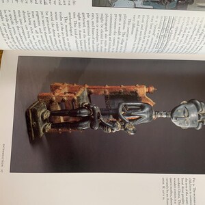 May include: A wooden sculpture of a figure with a large head and elongated body, holding a smaller figure. The sculpture is dark brown and black, with intricate details. The figure is mounted on a rectangular base. The sculpture is displayed on a page with text.