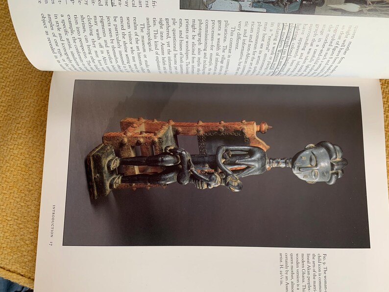May include: A wooden sculpture of a figure with a large head and elongated body, holding a smaller figure. The sculpture is dark brown and black, with intricate details. The figure is mounted on a rectangular base. The sculpture is displayed on a page with text.