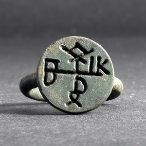 Könnte beinhalten: Ein bronzener Ring mit einer kreisförmigen Oberfläche, die ein eingraviertes Design aufweist. Der Ring hat eine dunkle, gealterte Patina, was darauf hindeutet, dass es sich um ein antikes oder Vintage-Stück handelt. Das Design auf der Oberfläche scheint stilisierte Buchstaben oder Symbole zu sein.