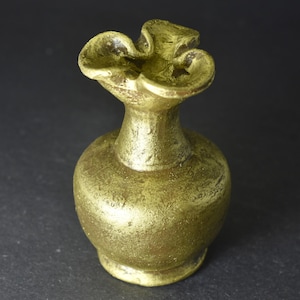 Puede incluir: Un pequeño jarrón dorado con la parte superior con volantes. El jarrón tiene un cuerpo redondeado y un cuello delgado, con una superficie texturizada. El diseño general es simple y elegante, adecuado para pequeños arreglos florales o decorativos.