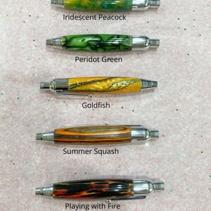 May include: Five pens with chrome hardware and patterned barrels labeled Iridescent Peacock, Peridot Green, Goldfish, Summer Squash, and Playing with Fire. Each pen features a unique color scheme including swirls of blue, green, yellow, wood grain, and black with orange accents.