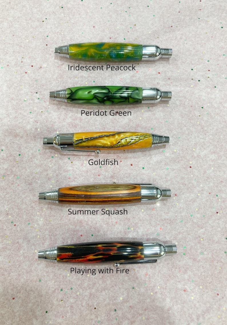 May include: Five pens with chrome hardware and patterned barrels labeled Iridescent Peacock, Peridot Green, Goldfish, Summer Squash, and Playing with Fire. Each pen features a unique color scheme including swirls of blue, green, yellow, wood grain, and black with orange accents.