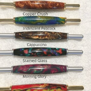 May include: Six cylindrical pen blanks with metal hardware, labeled Copper Crush in brown, Iridescent Peacock in blue and green, Cappuccino in brown, Stained Glass in multi-color, Morning Glory in pink and green, and Midnight Bonfire in red and yellow.
