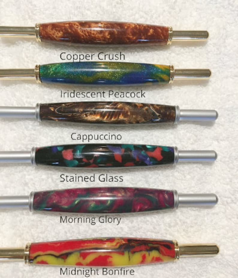 May include: Six cylindrical pen blanks with metal hardware, labeled Copper Crush in brown, Iridescent Peacock in blue and green, Cappuccino in brown, Stained Glass in multi-color, Morning Glory in pink and green, and Midnight Bonfire in red and yellow.