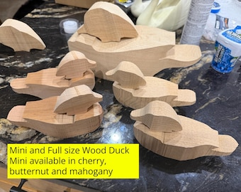 May include: A collection of unfinished light-colored wooden duck figurines in various sizes, including one large duck and several smaller duck shapes. The text on the image reads: Mini and Full size Wood Duck Mini available in cherry, butternut and mahogany.